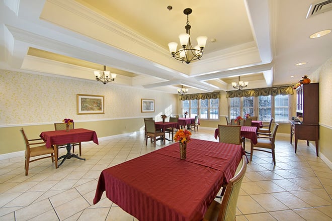 A spacious dining area with tables covered in red tablecloths, illuminated by elegant chandeliers and natural light from large windows.