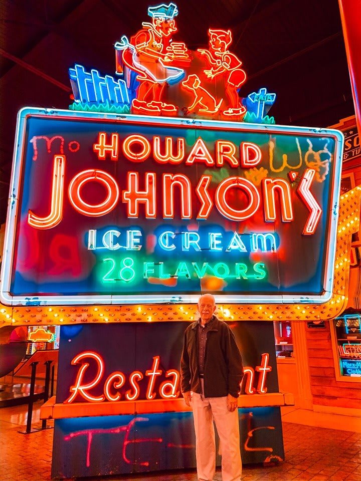 A person standing in front of a brightly lit Howard Johnson's neon sign advertising ice cream and a restaurant.