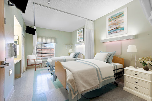 A clean and modern hospital patient room featuring two beds, light green walls, and natural light.