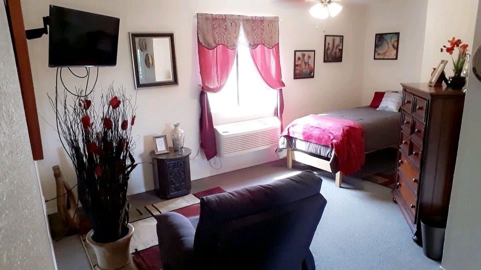 A cozy bedroom with red accents, featuring a bed, chair, window with curtains, and decorative items.
