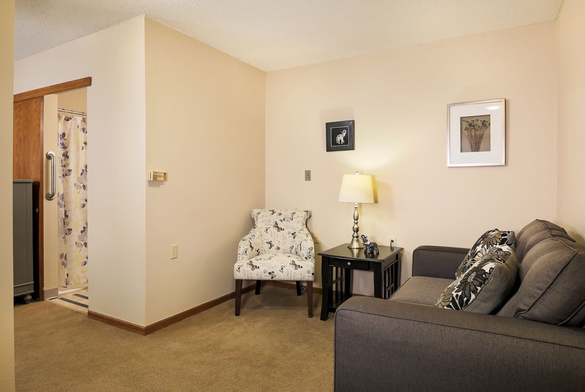 A cozy living room with a floral chair, sofa, lamp on a side table, and artwork on the walls.