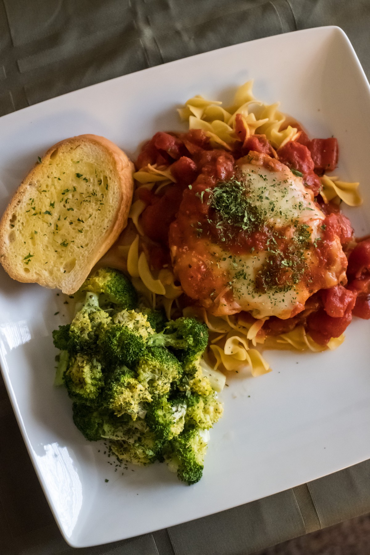 A delicious dinner plate featuring pasta with chicken in tomato sauce, steamed broccoli, and a slice of garlic bread.