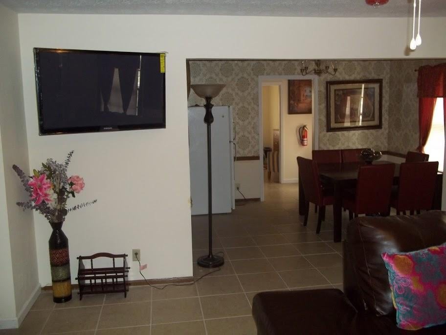 A cozy living room with a mounted TV, floor lamp, and dining area in the background.
