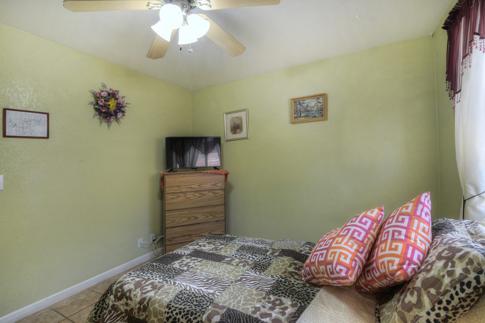 A cozy bedroom interior featuring a bed with patterned bedding, a wooden dresser topped with a television, and a ceiling fan overhead.