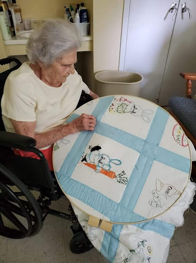 An elderly woman in a wheelchair is focused on her embroidery work at a table.