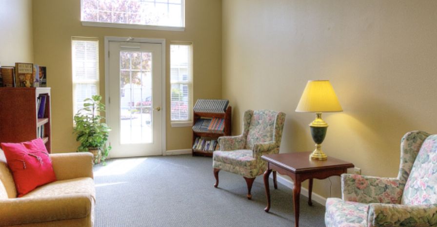 A cozy living room corner with a chair, table, lamp, and bookshelf near a bright window.