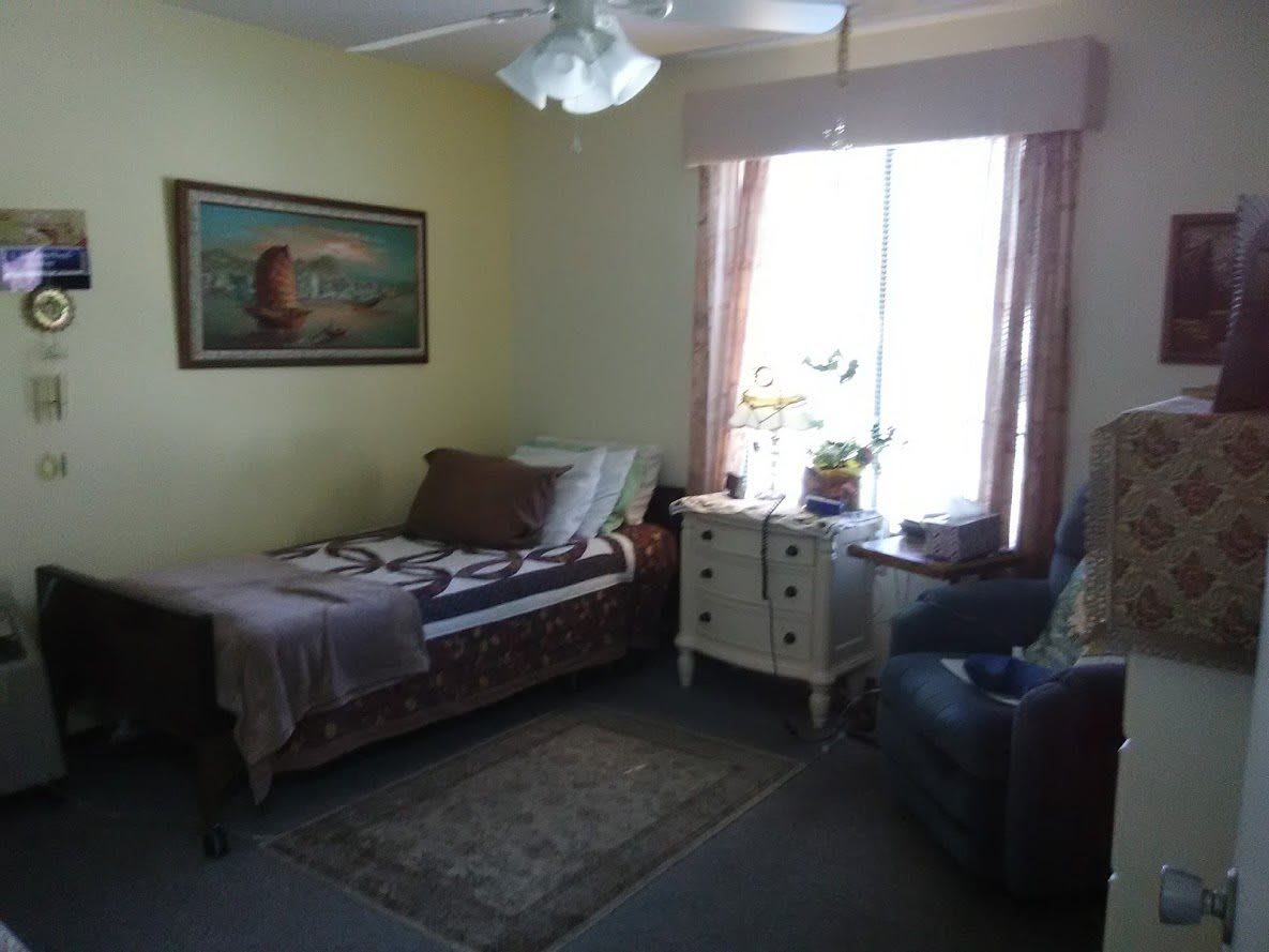 A cozy bedroom featuring a single bed, an armchair, a dresser by the window with curtains, and a painting on the wall.