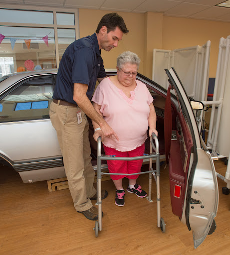 A caregiver assists an elderly woman using a walker to exit a car in a healthcare setting.