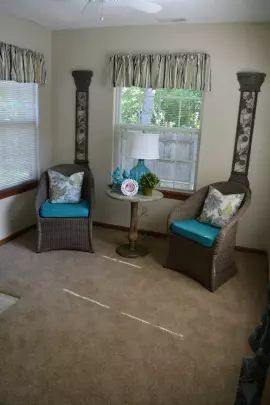 A cozy sitting area with two wicker chairs and a small round table between them, placed near windows with patterned curtains.