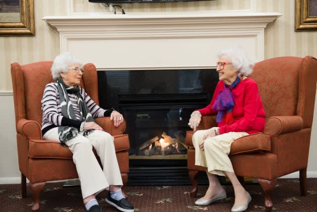 Two elderly women sitting by a fireplace having a conversation indoors.
