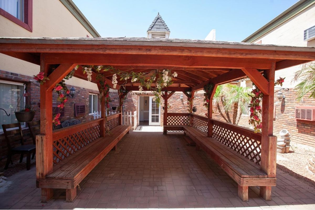 A wooden gazebo with bench seating, featuring lattice designs, located between brick buildings.