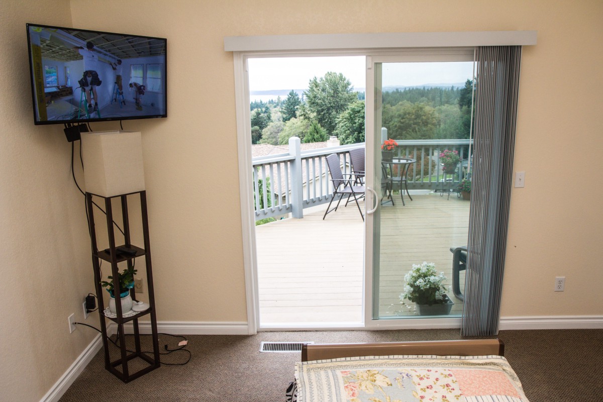 A cozy bedroom with a sliding glass door leading to a balcony overlooking nature, featuring a mounted television and simple decor.