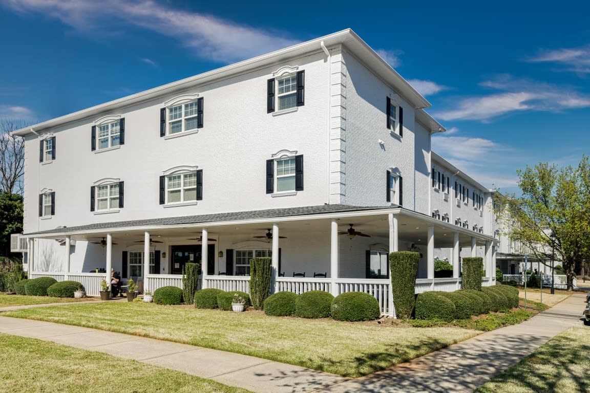 A three-story white building with black shutters and a wraparound porch, surrounded by neatly maintained landscaping.