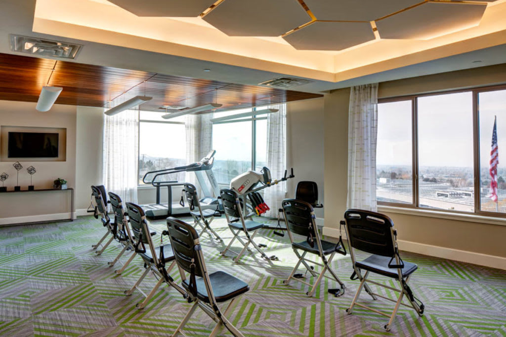 A bright exercise room with chairs arranged and a treadmill near large windows.