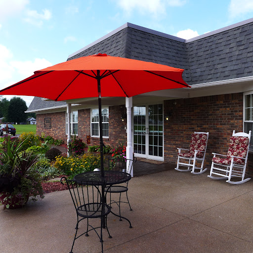A cozy outdoor patio featuring a red umbrella over a black table and chairs, surrounded by greenery.