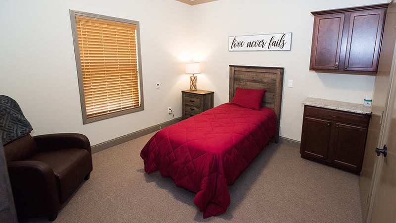 A cozy bedroom with a single bed featuring red bedding, a nightstand with a lamp, and window blinds.