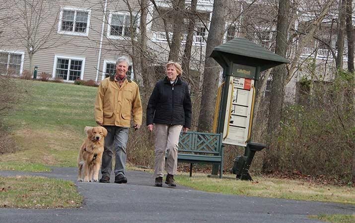 Two people walking a dog on a path near trees and a building.