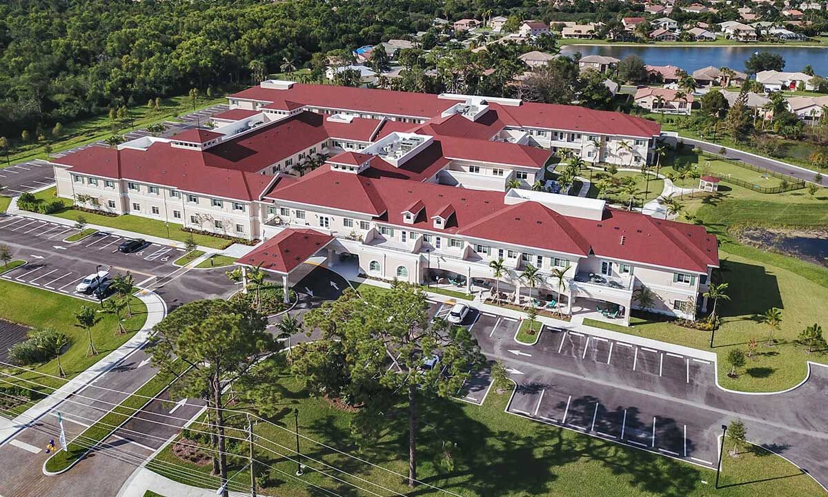 Aerial view of a large building with a red roof surrounded by greenery and parking areas.