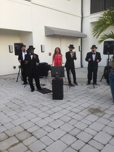 A lively outdoor performance featuring a band dressed in black suits and hats, accompanied by a female singer in a vibrant pink outfit.