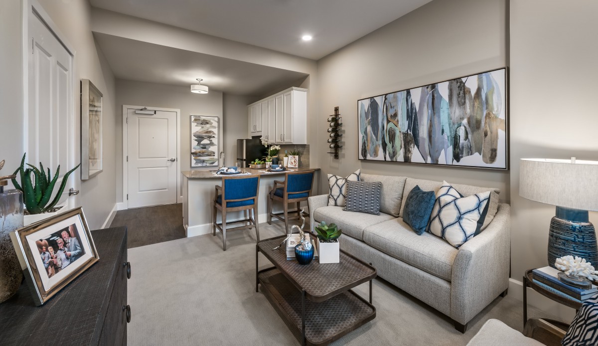 A modern apartment living room with a sofa, coffee table, and stylish artwork on the wall. The open kitchen area is visible in the background.