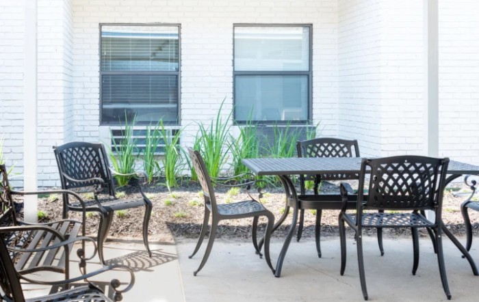 A modern patio with a set of outdoor furniture, featuring a table and chairs, surrounded by plants and large windows.