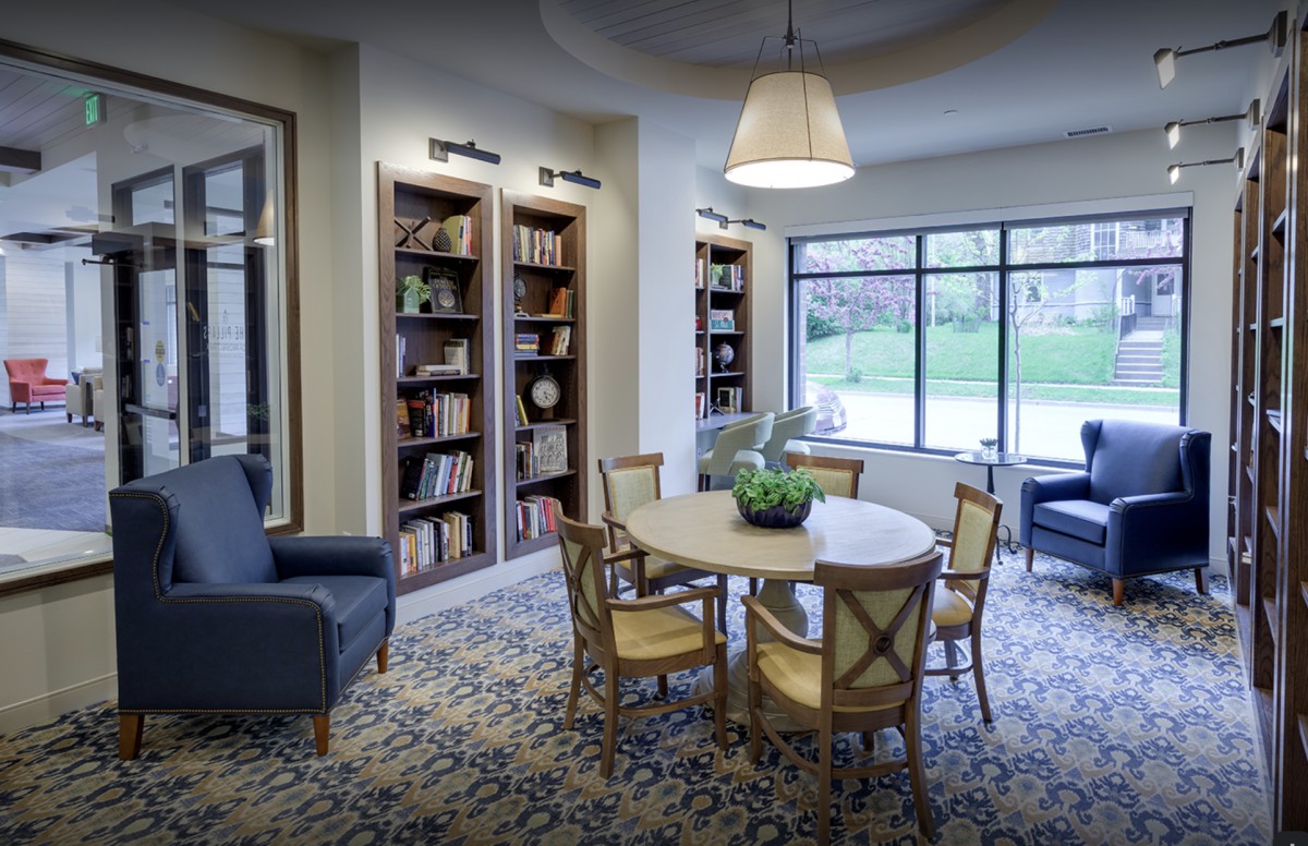 A cozy library reading area with bookshelves, a round table and chairs, illuminated by natural light from large windows.