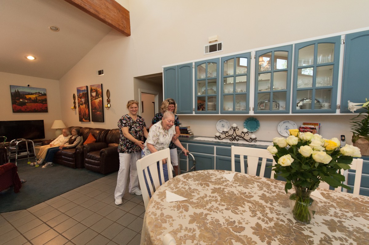 Two elderly individuals in a cozy kitchen, with one assisting the other as they walk.