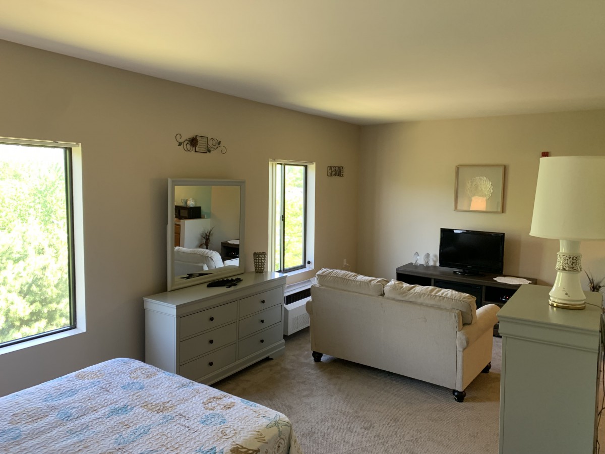 A cozy living space with a bed, dresser, sofa, and television, featuring neutral tones and natural light.