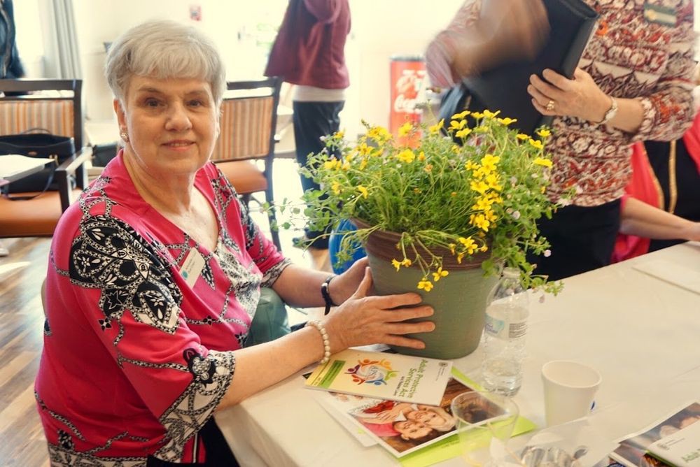 A woman sitting at a table holding a flower pot with yellow flowers during an indoor event.
