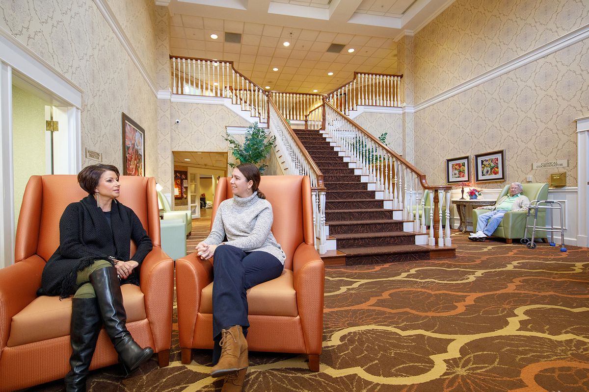 Two people conversing in a hotel lobby with a grand staircase in the background.