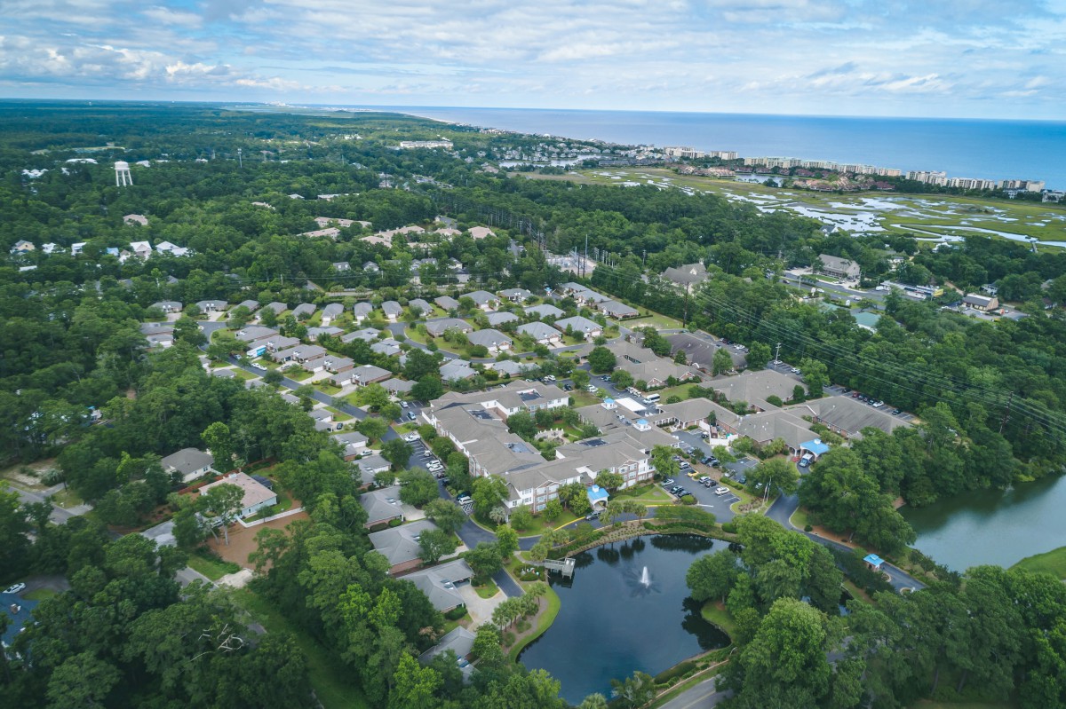 Aerial view of a residential neighborhood with houses, surrounded by greenery and a nearby lake, leading to a distant coastline.