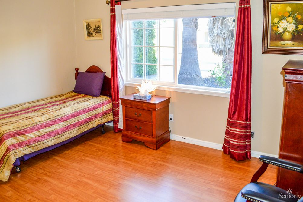 A cozy bedroom with a single bed, wooden floors, and a large window with red curtains.