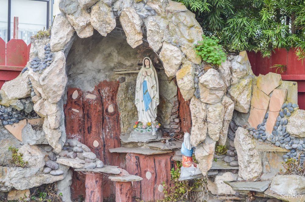 A religious grotto featuring a statue of the Virgin Mary, surrounded by stones and greenery.