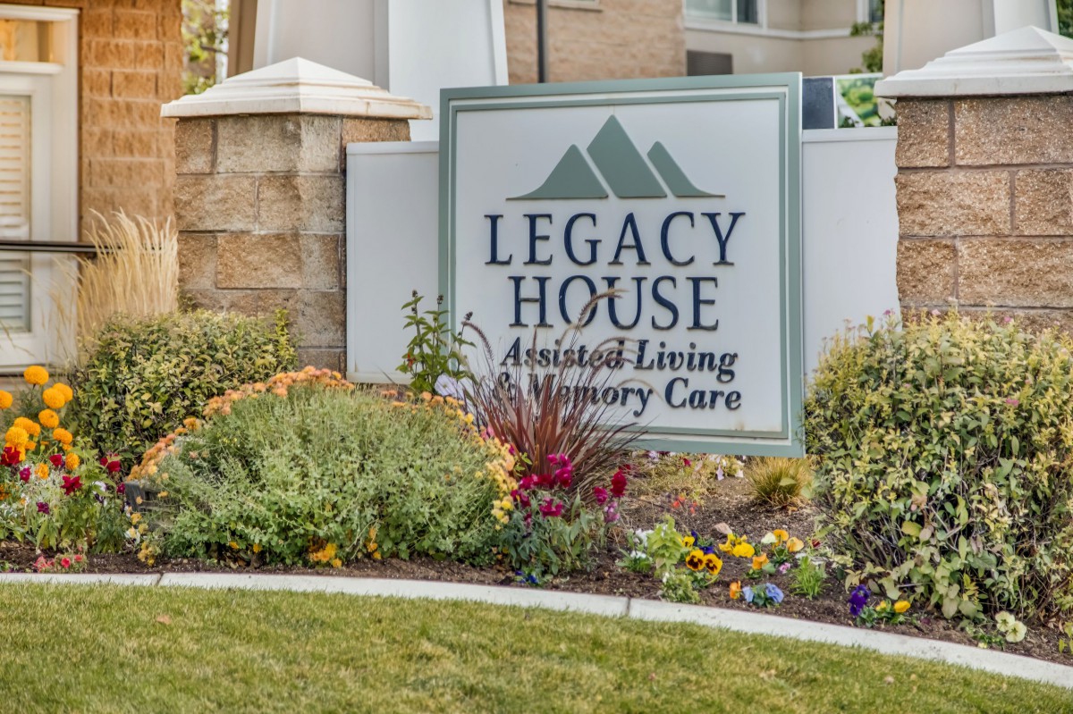 A sign for Legacy House, an assisted living and memory care facility, surrounded by landscaped greenery and flowers.