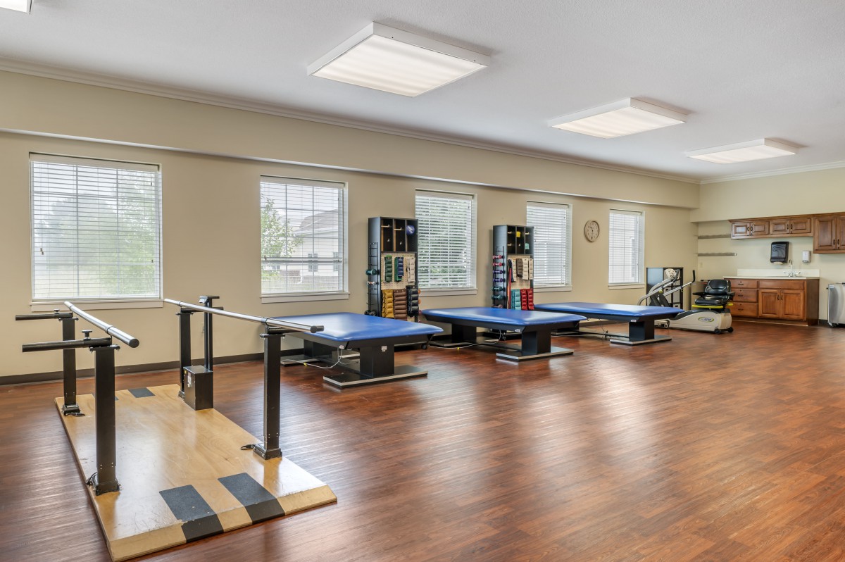 A spacious rehabilitation room with exercise equipment and wooden flooring, illuminated by natural light through large windows.