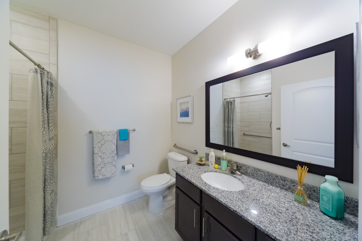 A modern bathroom featuring a granite countertop, sink, mirror, shower, and neatly hung towels.