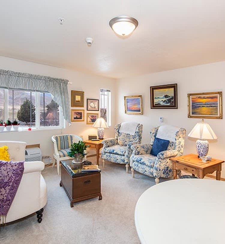 A cozy living room with floral-patterned armchairs, various artworks on the walls, and soft lighting from table lamps.