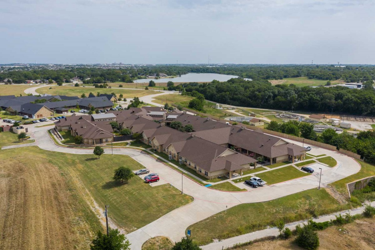 Aerial view of a residential area with multiple buildings, surrounded by grass and trees, featuring a parking lot.