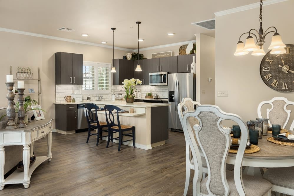 A modern kitchen with a dining area, featuring dark cabinets, stainless steel appliances, and pendant lighting.