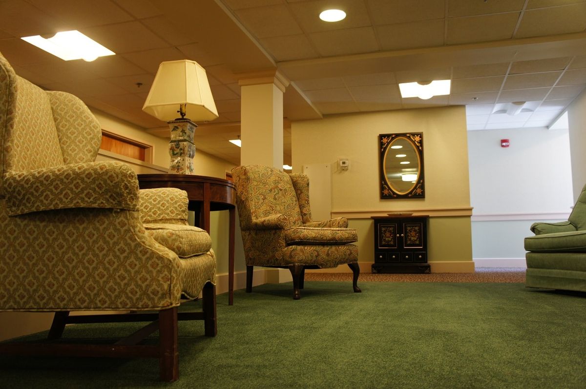 A cozy hallway featuring upholstered chairs, a carpeted floor, a table with a lamp, and a decorative mirror.