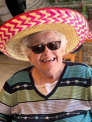 An elderly woman smiling, wearing sunglasses and a colorful sombrero.