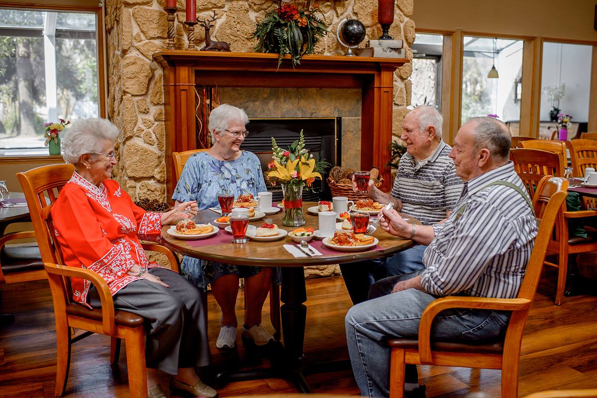 A group of elderly friends enjoys a meal together around a table in a cozy room with a fireplace.