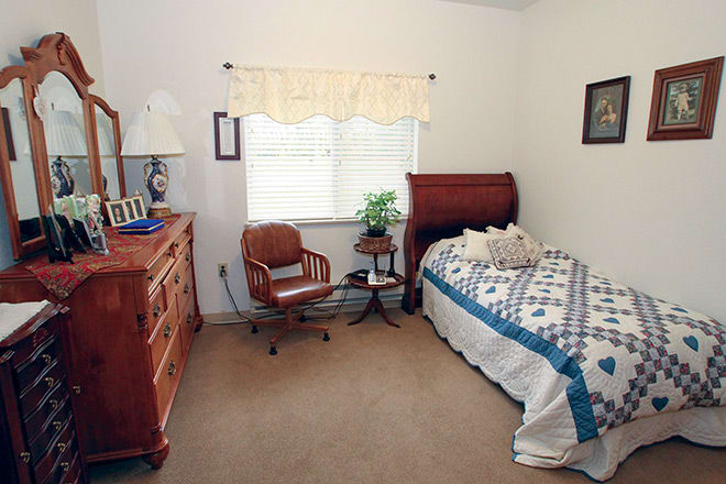 A cozy bedroom featuring a wooden bed, chair, and dresser with a quilted bedspread and a window with curtains.
