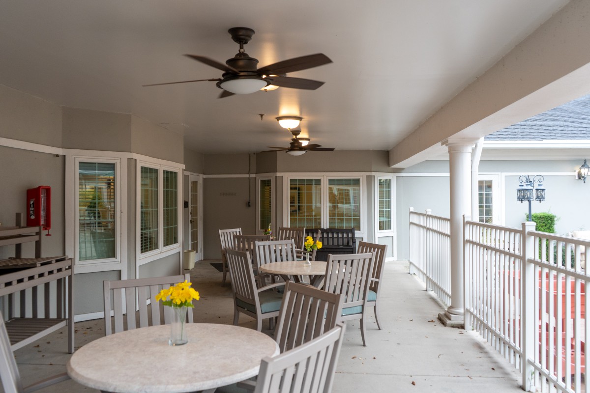 A covered patio with multiple tables and chairs, featuring ceiling fans and decorative flowers on each table.