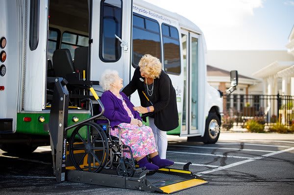 A caregiver assists an elderly woman in a wheelchair boarding a bus using a wheelchair lift.