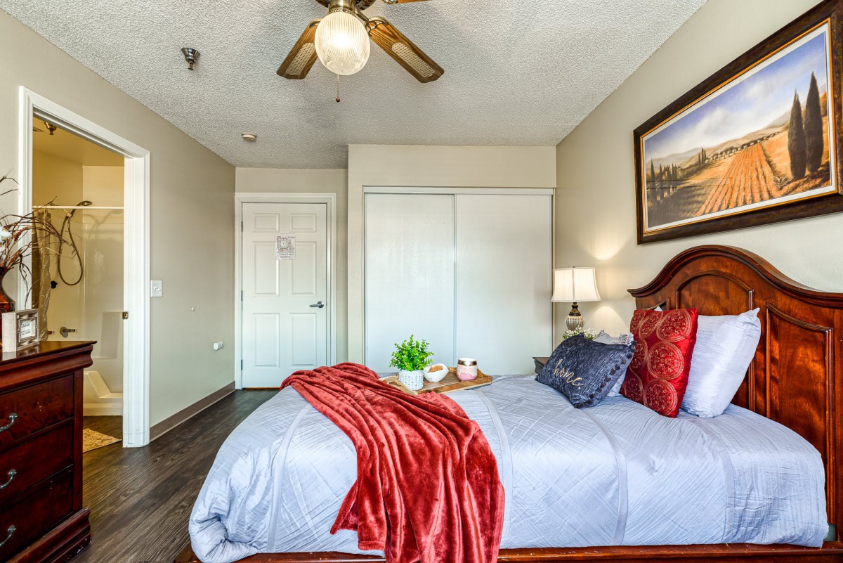 A cozy bedroom with a neatly made bed, decorative pillows, and a red throw blanket. The room features a ceiling fan, a bedside lamp, and artwork on the wall.