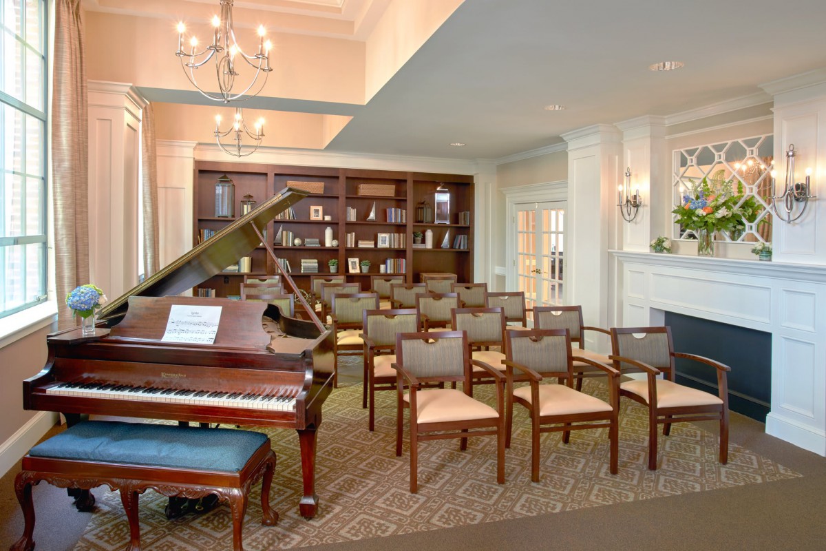 A cozy meeting room featuring a grand piano and neatly arranged chairs facing a bookshelf.