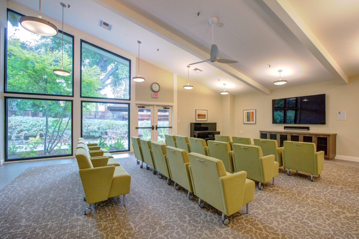 A well-lit conference room with multiple rows of green chairs facing a television and large windows.
