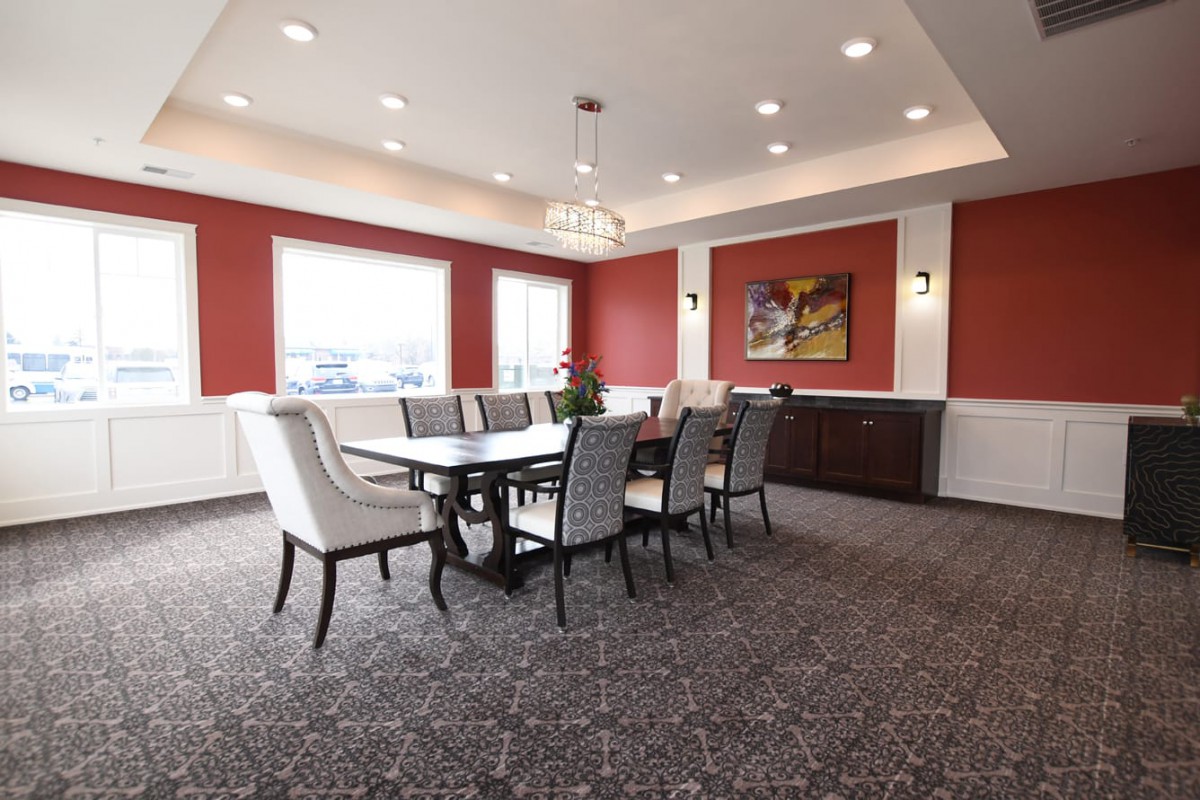 A modern dining room with a large table, surrounded by upholstered chairs, featuring red accent walls and elegant lighting.