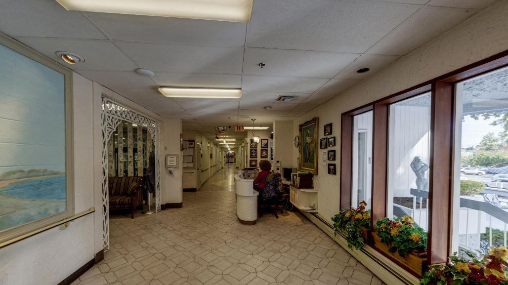 A well-lit hallway with a reception desk, seating area, and plants near the windows.